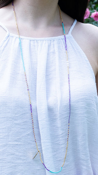 A woman wearing a colourful layered beaded necklace with turquoise, lilac, fuchsia, freshwater pearl, and gold accents, adding an elegant pop of colour to a light summer outfit with effortless style.